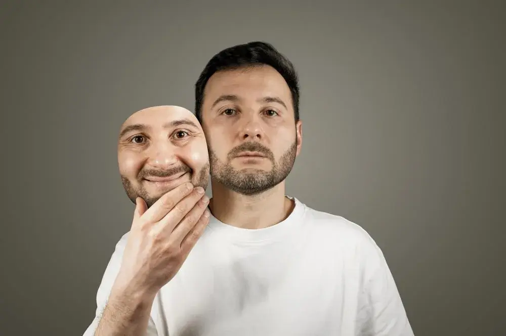 A man in a white shirt holds a smiling mask in front of his face, revealing a neutral expression underneath.