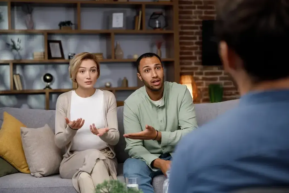 A woman and a man are sitting on a couch, gesturing and talking during couples therapy