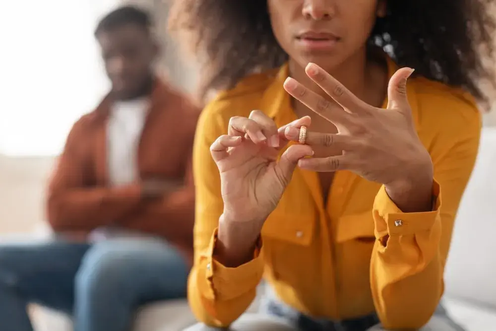 A woman sits on a couch removing a ring from her finger, while a man in the background sits with his arms crossed.