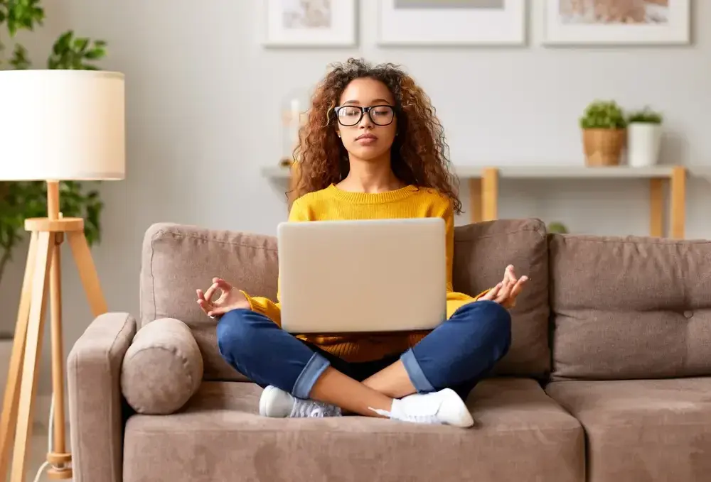 A person with curly hair and glasses sitting cross-legged on a couch with a laptop, eyes closed, hands resting in a meditative pose.