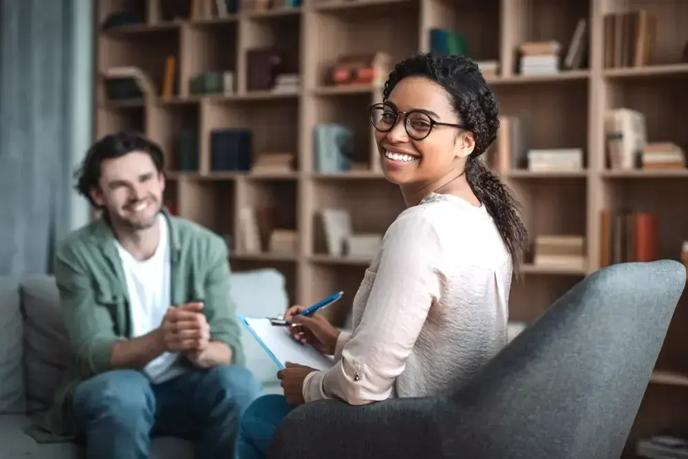 A woman with glasses taking notes while a man sits across from her on a couch, a bookshelf behind them.