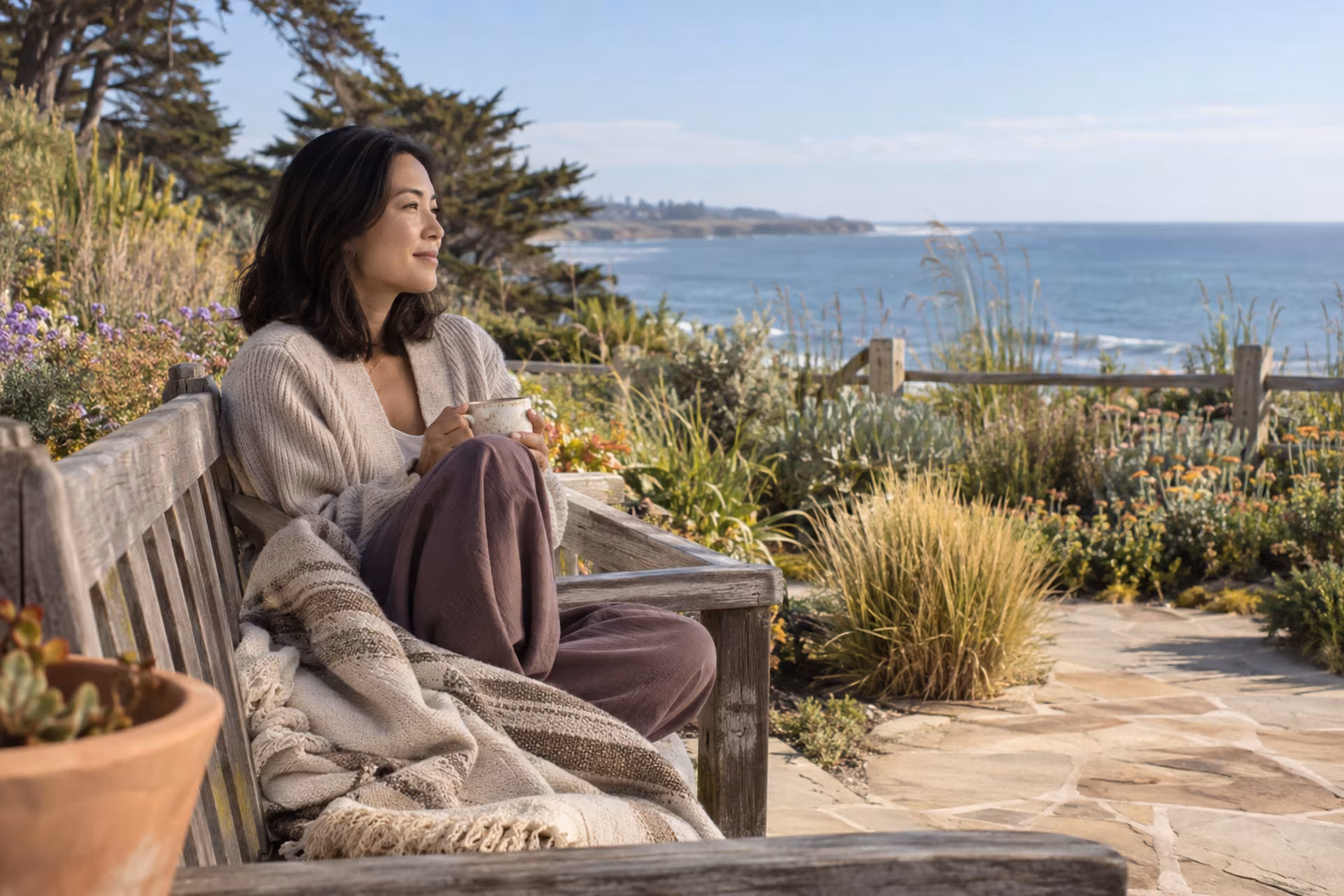 A woman in a cream cardigan sits on a wooden garden bench overlooking the coast, holding a mug during an emotional healing retreat.