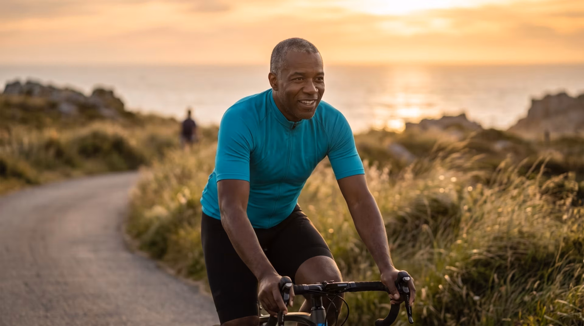 A man in his fifties cycling along a coastal path at sunrise, smiling and riding with easy energy.