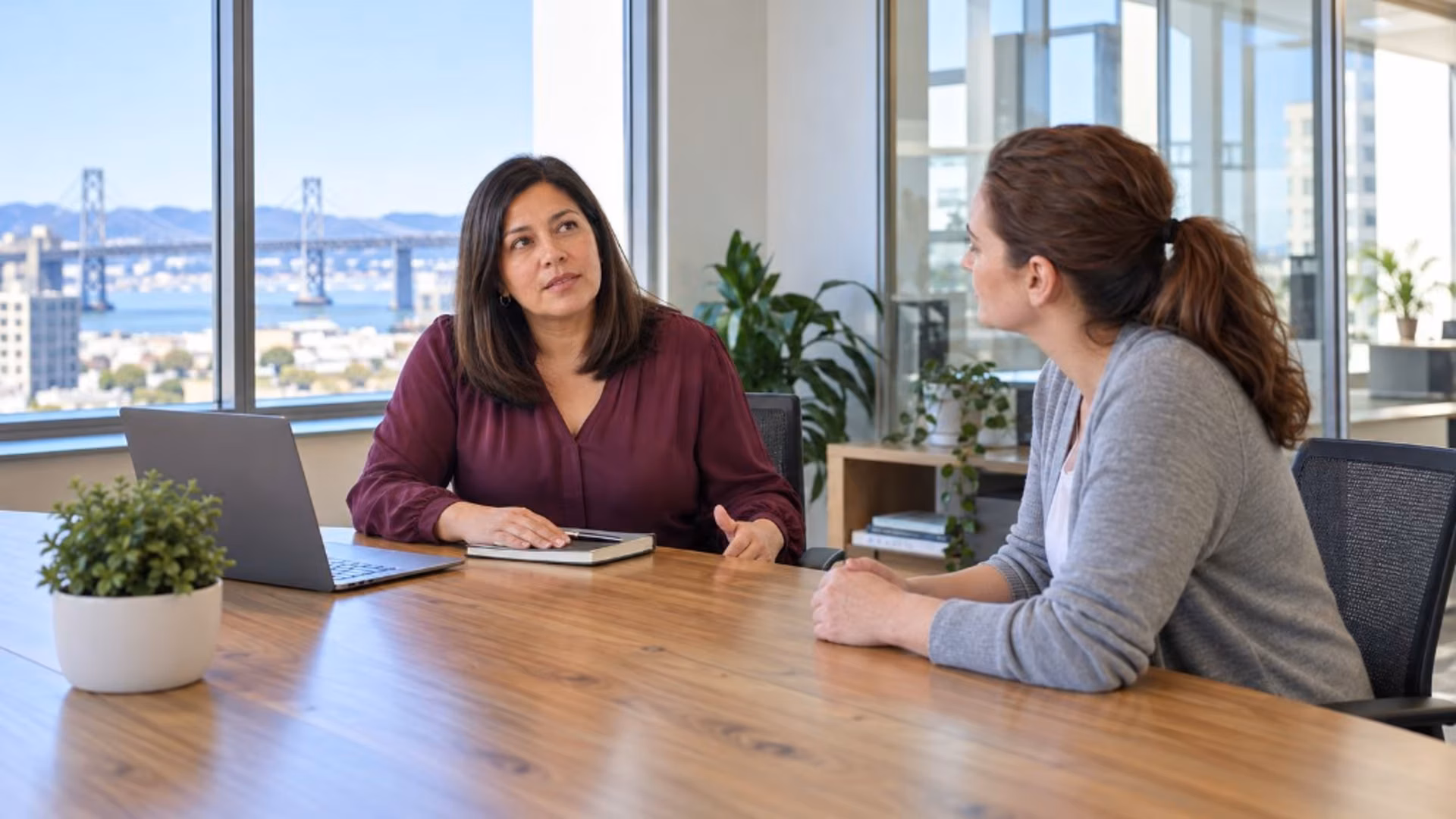 Two women sit at a wood conference table in a high-rise, the Bay Bridge behind them, one in a burgundy blouse talking while the other in a gray cardigan listens