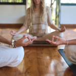 In a serene indoor setting, three people are sitting cross-legged on a wooden floor, meditating with eyes closed. This tranquil scene captures the essence of a mental health retreat, fostering peace and rejuvenation for all who partake.