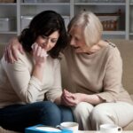 Two women sit on a couch; one is finding her way through ambiguous grief, while the other offers comfort, tissue in hand. Two cups and a tissue box rest on the table before them.