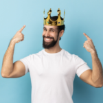 Man with a beard wearing a gold crown, smiling and pointing at the crown, standing against a blue background, exuding an air of confidence that hints playfully at Narcissistic Personality Disorder.