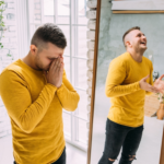 A person in a yellow sweater stands by a mirror, appearing distressed, while their reflection shows them laughing—an evocative portrayal of bipolar disorder's dual nature. A potted plant and large window create a serene backdrop, emphasizing the contrast in emotions.