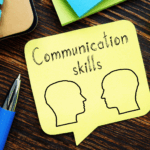 A yellow sticky note with "Communication skills" and "Understanding Needs" written above two outline drawings of human heads, placed on a wooden desk with a pen and office supplies nearby.