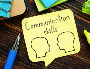 A yellow sticky note with "Communication skills" and "Understanding Needs" written above two outline drawings of human heads, placed on a wooden desk with a pen and office supplies nearby.