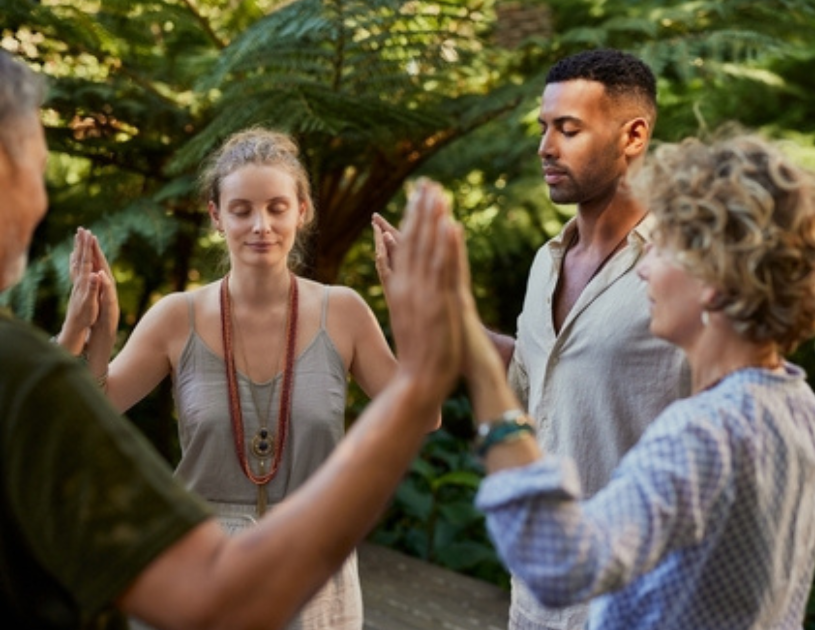 Four adults stand in a circle outdoors with eyes closed, palms touching, participating in a mindfulness or group meditation activity inspired by practices like CBT therapy.