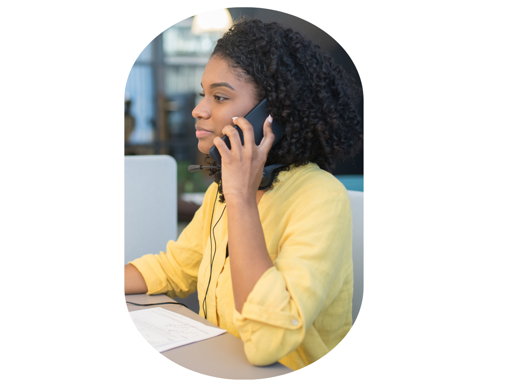 A woman wearing a yellow shirt is sitting at a desk, talking on the phone with papers and a laptop in front of her, perhaps searching for a couples spa retreat near me.