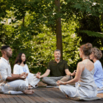 Five people sit cross-legged in a circle on a wooden deck, meditating together in a lush, green forest—an ideal scene for a meditation retreat at one of 2025’s top destinations.