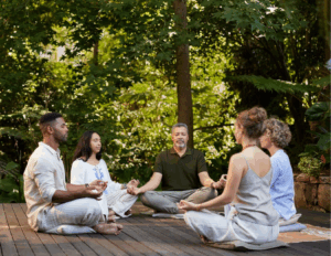 Five people sit cross-legged in a circle on a wooden deck, meditating together in a lush, green forest—an ideal scene for a meditation retreat at one of 2025’s top destinations.