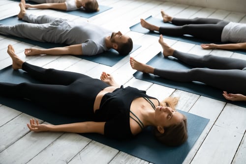 Several people lie on yoga mats in a spacious room, practicing a relaxation pose often used in CBT therapy, with their eyes closed and arms at their sides.