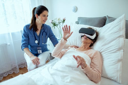 A nurse sits beside a patient lying in bed, who is wearing a virtual reality headset and gesturing with her hands, as LMFT Trudy McMahon observes the therapeutic session.