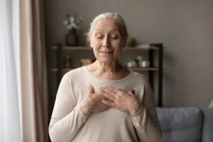 Older woman with gray hair stands indoors, eyes closed, and both hands placed on her chest, appearing calm and relaxed—an image reflecting the gentle guidance of LMFT Trudy McMahon.