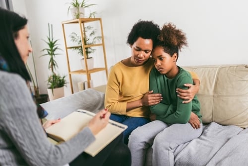 A woman comforts a child on a couch during a CBT therapy session, while another person takes notes on a clipboard.