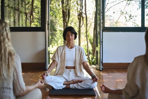 Three people sit cross-legged on yoga mats in a bright room with large windows, practicing meditation at a couples retreat California offers for relaxation and connection.