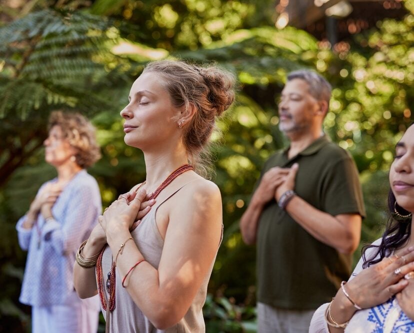 A group of people stand outdoors with eyes closed, hands on their chests, meditating or practicing mindfulness during wellness retreats in a lush, green setting.