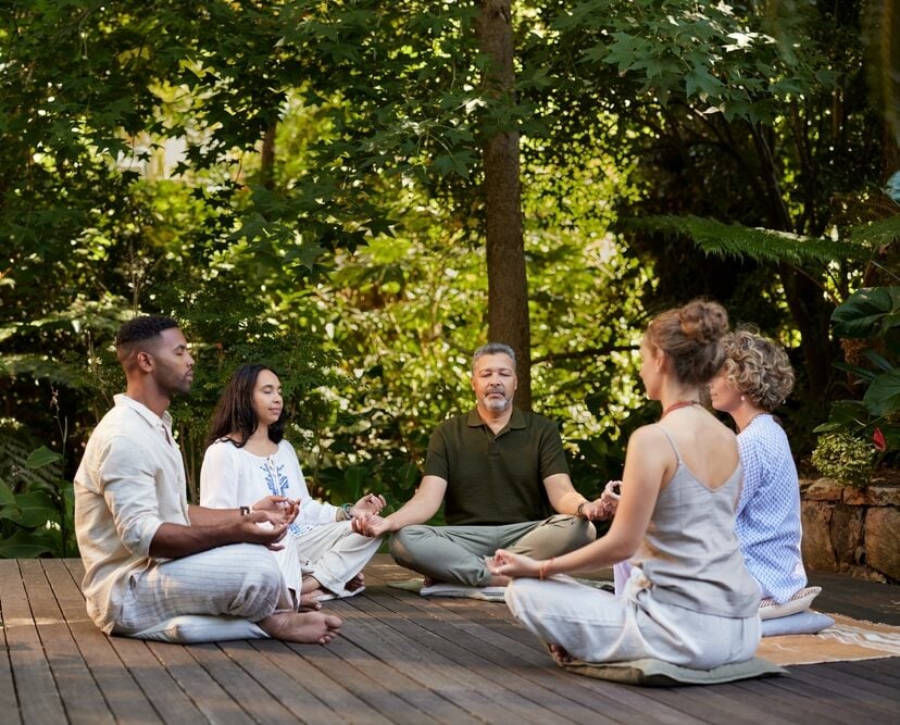 Five people sit cross-legged on a wooden deck outdoors, meditating together in a lush, green garden setting—an inspiring glimpse into the serenity of wellness retreats.