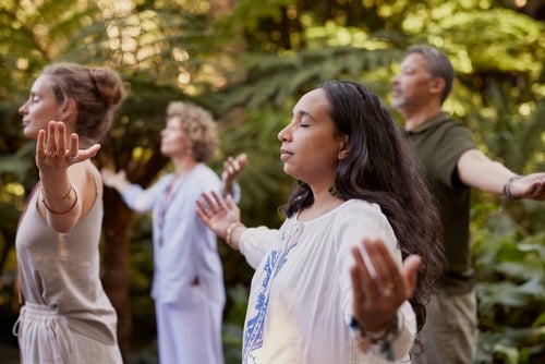 Four adults stand outdoors with eyes closed and arms raised, appearing to engage in a meditative practice—an inspiring scene for those seeking a couple retreat near me surrounded by peaceful greenery.