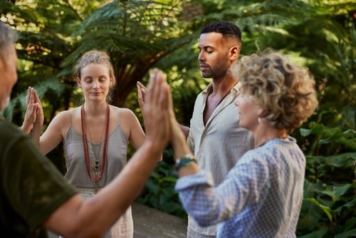 Four adults stand in a circle outdoors, eyes closed and palms pressed together in pairs, surrounded by lush greenery—an ideal moment from a couples retreat California experience.