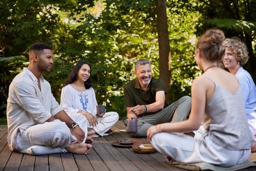 Five adults sit on a wooden deck outdoors, talking and smiling, surrounded by greenery. Some hold mugs and a teapot sits in the center—perfect inspiration for a cozy couples retreat near me.