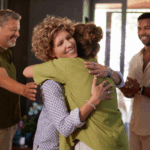 Two women are hugging and smiling indoors, while two men stand nearby, watching and clapping—celebrating their mental health retreat preparation and the supportive journey ahead.