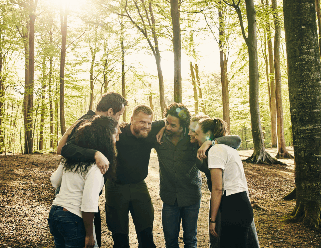 Five people stand in a circle in a sunlit California forest, smiling and leaning together with arms around each other's shoulders at a mental health retreat in California.
