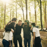 Five people stand in a circle in a sunlit California forest, smiling and leaning together with arms around each other's shoulders at a mental health retreat in California.