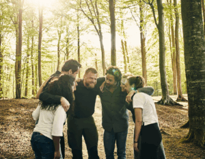 Five people stand in a circle in a sunlit California forest, smiling and leaning together with arms around each other's shoulders at a mental health retreat in California.