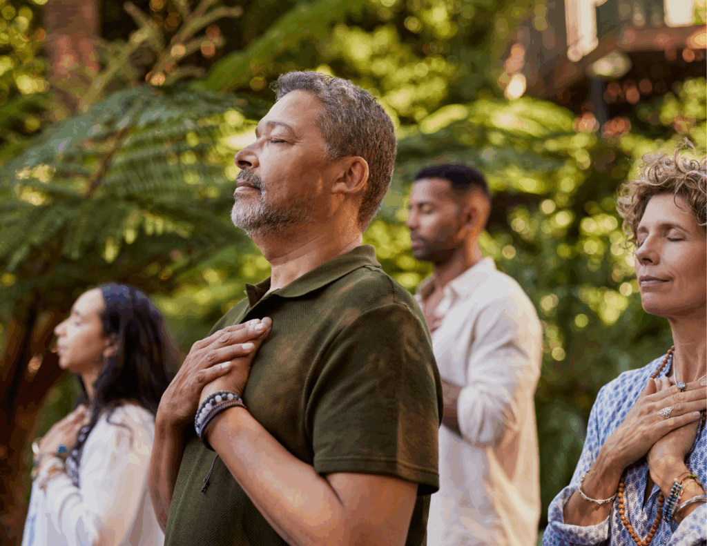 Four people stand outdoors with eyes closed and hands on their chests, appearing calm and focused, surrounded by greenery—a peaceful moment at a mental health retreat.