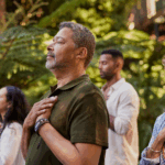 Four people stand outdoors with eyes closed and hands on their chests, appearing calm and focused, surrounded by greenery—a peaceful moment at a mental health retreat.