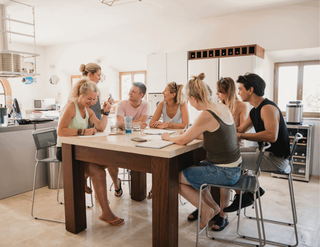 Seven people sit around a kitchen island engaged in conversation, writing, and drinking water in a bright, modern kitchen at a Behavioral Health Retreat near me.