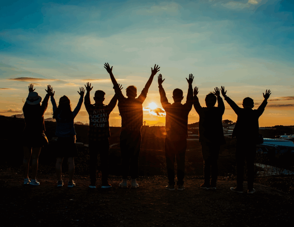 A group of seven people stands on a hill at sunset, arms raised in unity, silhouetted against the sky—capturing a moment of wellness and togetherness reminiscent of Collective Retreats at Bay Area CBT Center.