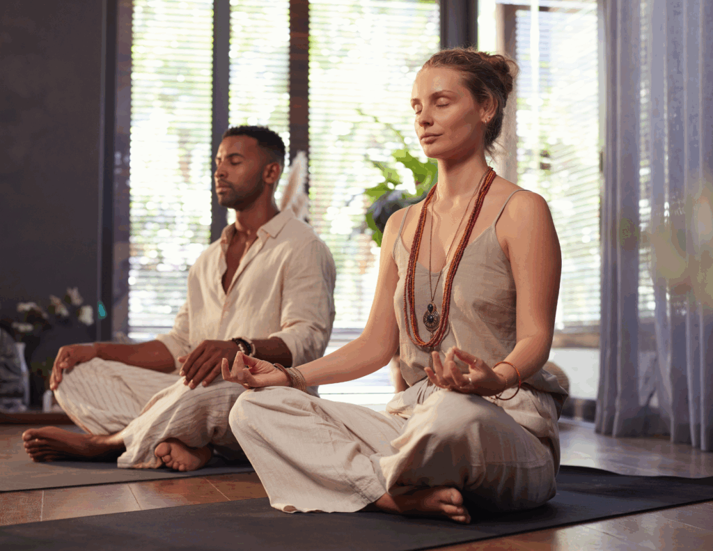 Two people sit cross-legged on yoga mats indoors, meditating with eyes closed and hands resting on their knees in a sunlit room—an ideal setting for couples retreats focused on strengthening relationship bonds.