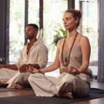 Two people sit cross-legged on yoga mats indoors, meditating with eyes closed and hands resting on their knees in a sunlit room—an ideal setting for couples retreats focused on strengthening relationship bonds.