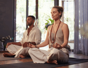 Two people sit cross-legged on yoga mats indoors, meditating with eyes closed and hands resting on their knees in a sunlit room—an ideal setting for couples retreats focused on strengthening relationship bonds.