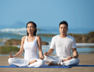 Two people sit cross-legged on yoga mats at the beach, meditating with closed eyes and relaxed expressions, dressed in white clothing. The serene Bay Area ocean and rocks provide a calming backdrop, perfect for couples retreat therapy.