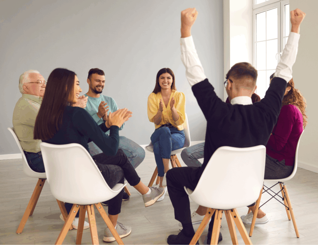 A group of six people sit in a circle on chairs indoors, clapping and smiling as one person raises both arms in celebration during a group therapy online support group for bipolar disorder near me.