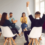 A group of six people sit in a circle on chairs indoors, clapping and smiling as one person raises both arms in celebration during a group therapy online support group for bipolar disorder near me.