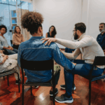A group of people sits in a circle on chairs in a bright room, highlighting the benefits of support groups, as one man offers comfort by placing his hand on another's shoulder.