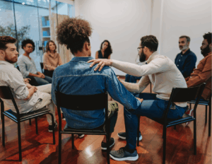 A group of people sits in a circle on chairs in a bright room, highlighting the benefits of support groups, as one man offers comfort by placing his hand on another's shoulder.