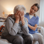 An elderly woman sits with her head in her hands while a caregiver in scrubs offers comfort, gently supporting her shoulder—an intimate moment that reflects the importance of grief counseling and emotional healing.