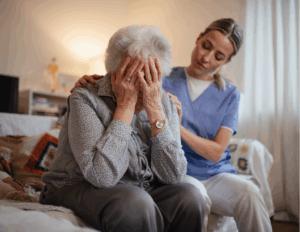 An elderly woman sits with her head in her hands while a caregiver in scrubs offers comfort, gently supporting her shoulder—an intimate moment that reflects the importance of grief counseling and emotional healing.