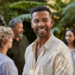 A man in a light shirt smiles in the foreground outdoors, radiating positivity, while three people converse amid greenery—a serene scene reminiscent of a mental health retreat.