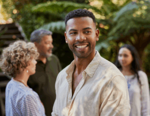 A man in a light shirt smiles in the foreground outdoors, radiating positivity, while three people converse amid greenery—a serene scene reminiscent of a mental health retreat.