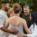 Four adults stand outside in a circle, holding each other in a group embrace, surrounded by greenery—capturing the sense of connection and well-being found at a Bay Area CBT Center mental health retreat.