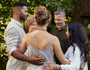 Four adults stand outside in a circle, holding each other in a group embrace, surrounded by greenery—capturing the sense of connection and well-being found at a Bay Area CBT Center mental health retreat.